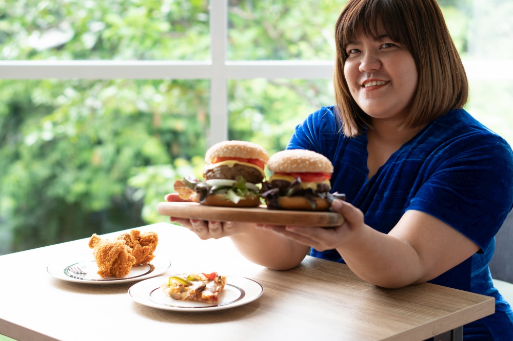 Hungry overweight woman holding hamburger on a wooden plate, Fried Chicken and Pizza on table, During work from home, gain weight problem. Concept of binge eating disorder (BED).