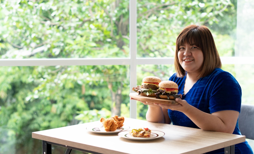 Hungry overweight woman holding hamburger on a wooden plate, Fried Chicken and Pizza on table, During work from home, gain weight problem. Concept of binge eating disorder (BED).
