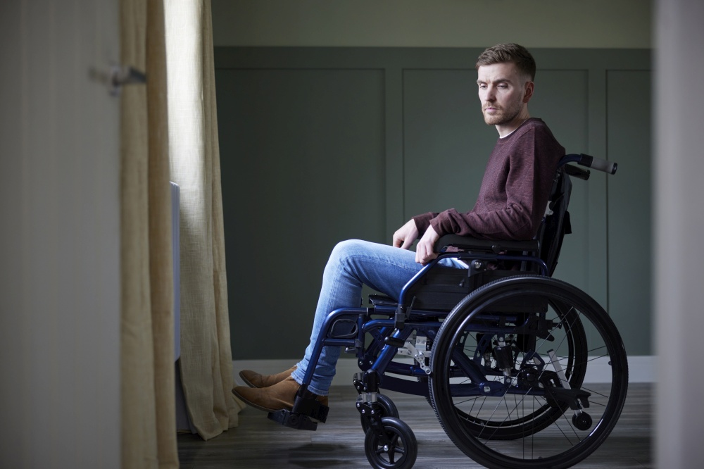 Depressed Young Man With Poor Mental Health In Wheelchair By Window At Home