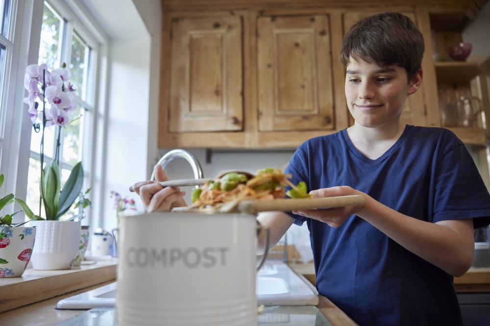 Boy In Kitchen Making Compost Scraping Vegetable Leftovers Into Bin