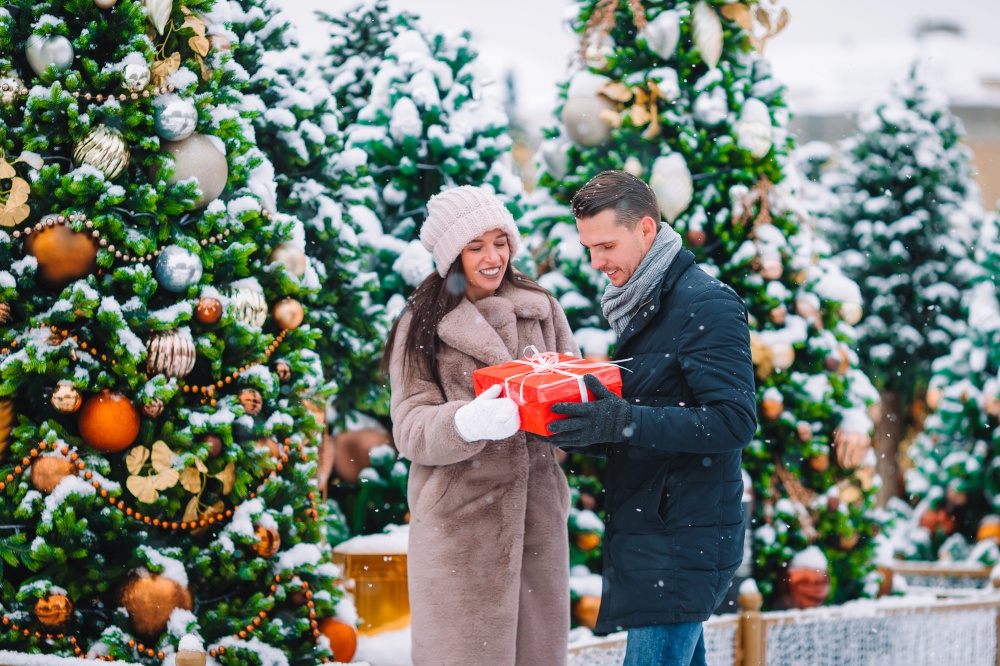 Young beautiful couple celebrating Christmas in the city street and giving gift to each other. Young beautiful cheerful couple celebrating Christmas in the city street