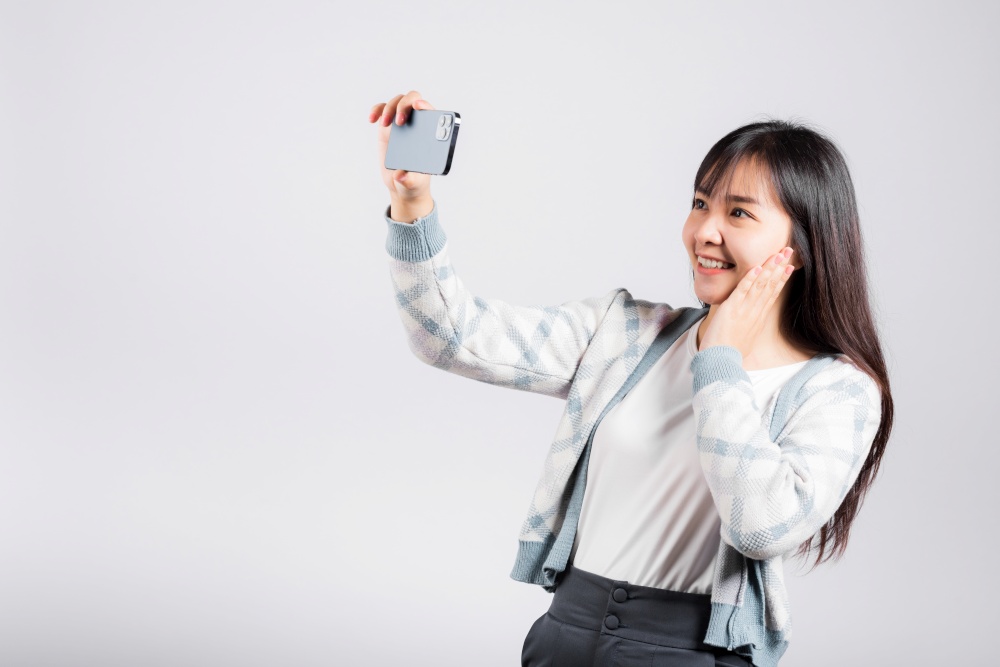 Woman excited holding smartphone to shooting selfie photo front camera studio shot isolated white background, happy young female smiling taking photography by mobile phone on mobile phone