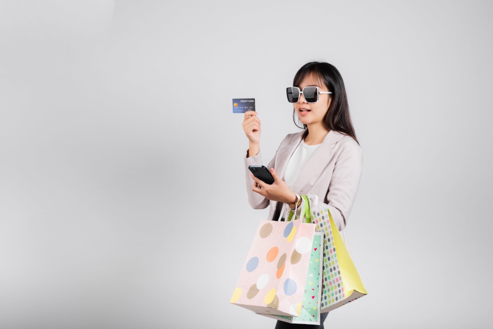 Woman with glasses shopper smile hold online shopping bags, smartphone and credit card for payment, Portrait excited happy Asian female sale mobile financial application isolated on white background