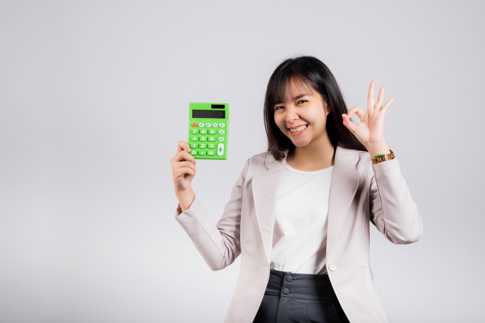 Tax day concept. Woman confident smiling holding electronic calculator and show OK gesture, excited happy Asian female studio shot isolated on white background, Account and finance counting income