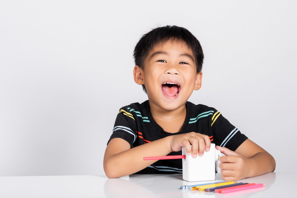 Little cute kid boy 5-6 years old smile using pencil sharpener while doing homework in studio shot isolated on white background, Asian children preschool sharpening color pencils, education concept