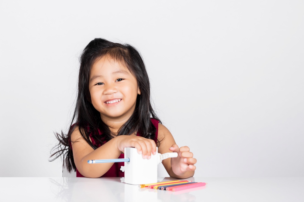 Little cute kid girl 3-4 years old smile using pencil sharpener while doing homework in studio shot isolated on white background, Asian children preschool sharpening color pencils, education concept