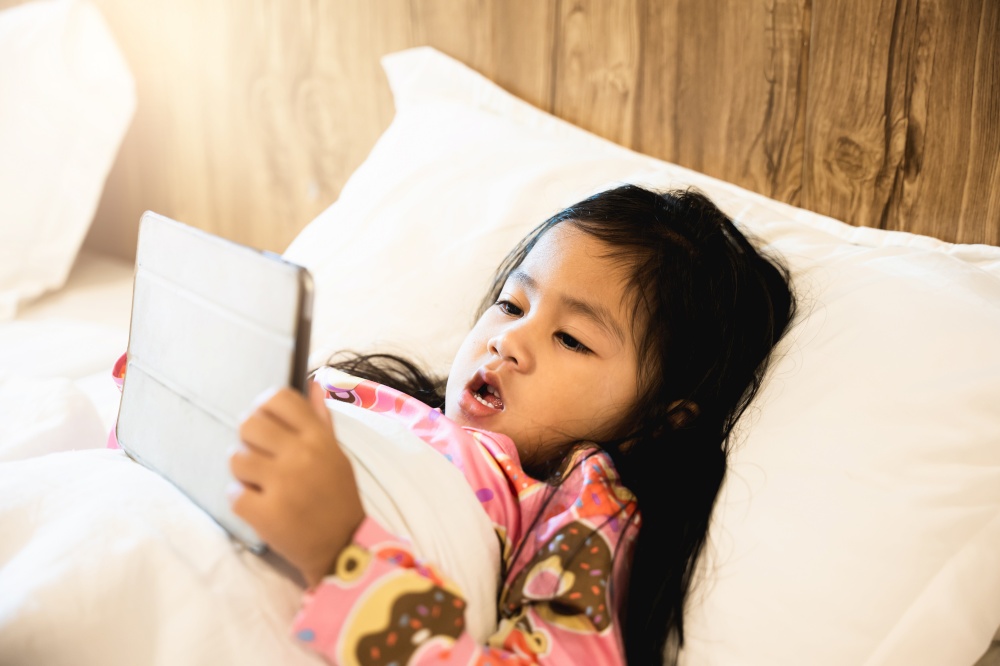 Little Asian girl playing digital tablet on bed addicted game and cartoon, Kid watching videos and cartoons online in bedroom at home, Fun online concept