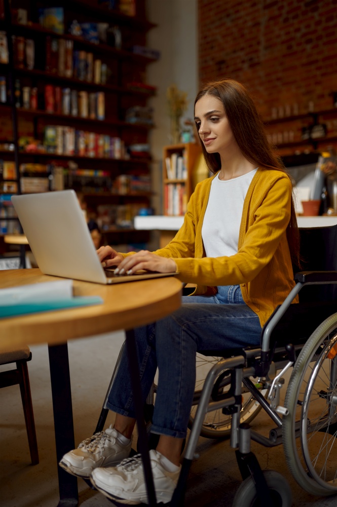Disabled female student in wheelchair using laptop, disability, bookshelf and university library interior on background. Handicapped young woman studying in college, paralyzed people get knowledge. Disabled female student in wheelchair using laptop