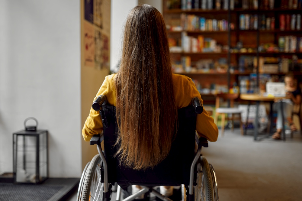 Disabled female student in wheelchair, back view, disability, bookshelf and university library interior on background. Handicapped young woman studying in college, paralyzed people get knowledge. Disabled female student in wheelchair, back view