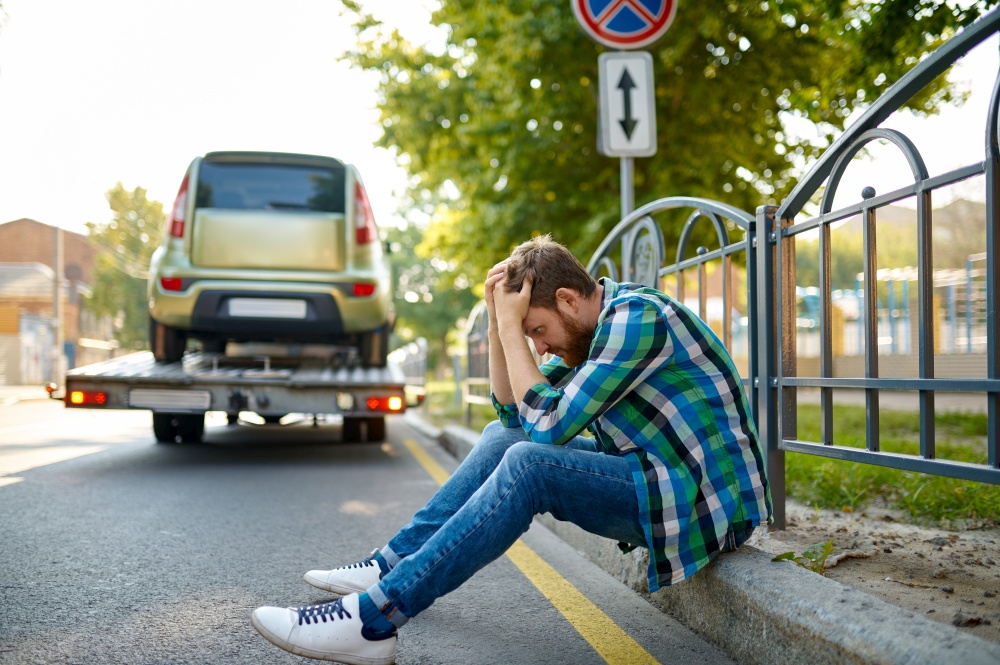 Unhappy man sitting on roadside holding his head during incorrect parked car evacuation by tow truck. Unhappy man and incorrect parked car evacuation