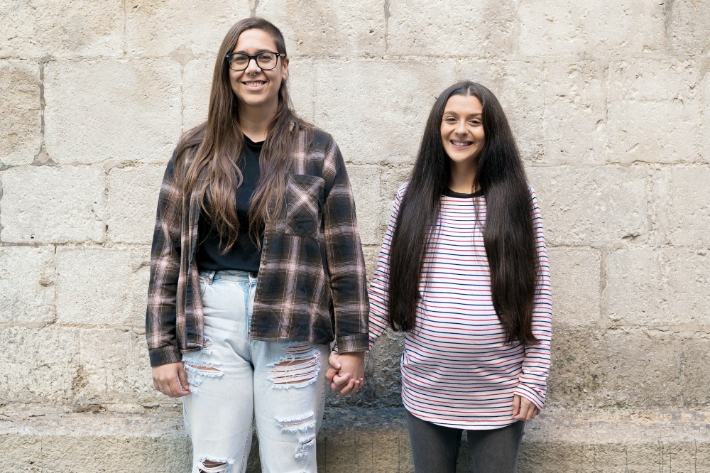 Two young lesbians holding each other and standing near a stone wall, smiling, happy. High quality photo. Two young lesbians holding each other and standing near a stone wall, smiling, happy.