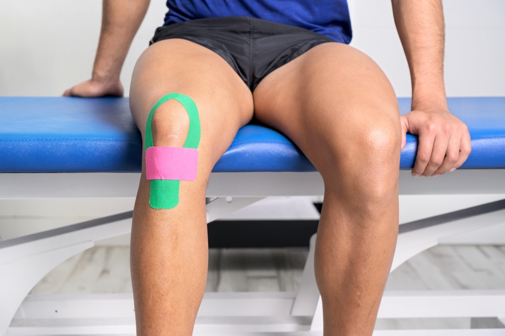 Young sportive male athlete treating injured knee with kinesio tape. Close up. Kinesiology, physical therapy, rehabilitation concept. High quality photo. Young sportive male athlete treating injured knee with kinesio tape. Close up. Kinesiology, physical therapy, rehabilitation concept.