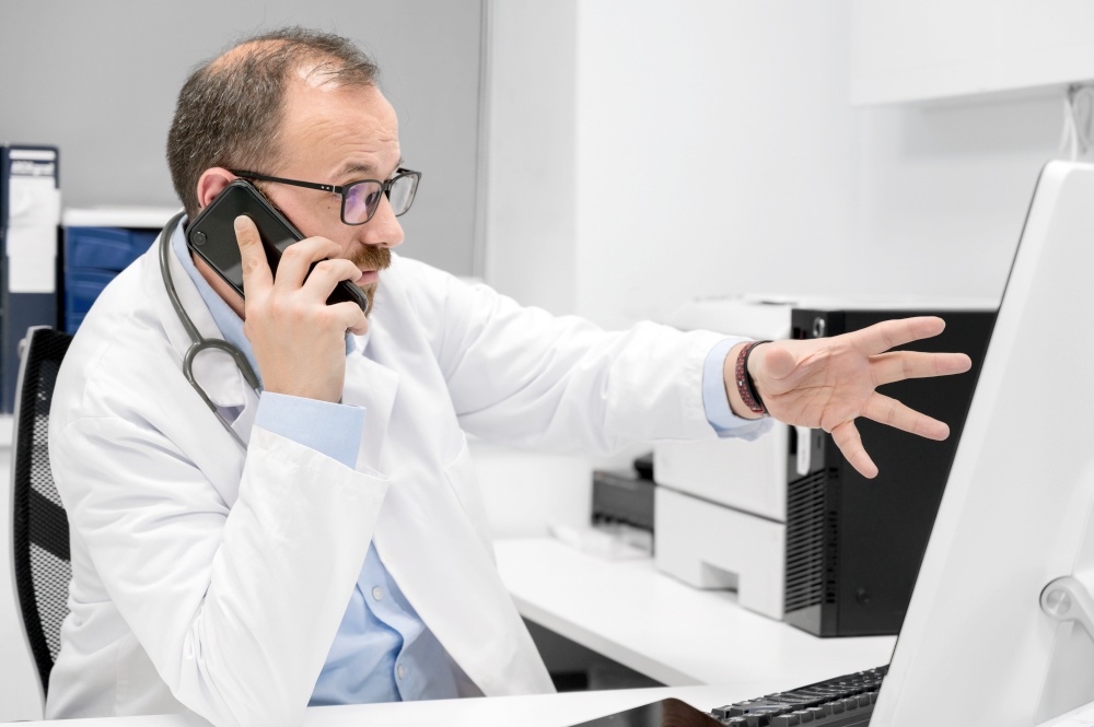Male doctor pointing with finger on desktop computer while talking on the phone, discussing treatment with colleague. High quality photo. Male doctor pointing with finger on desktop computer while talking on the phone, discussing treatment with colleague.