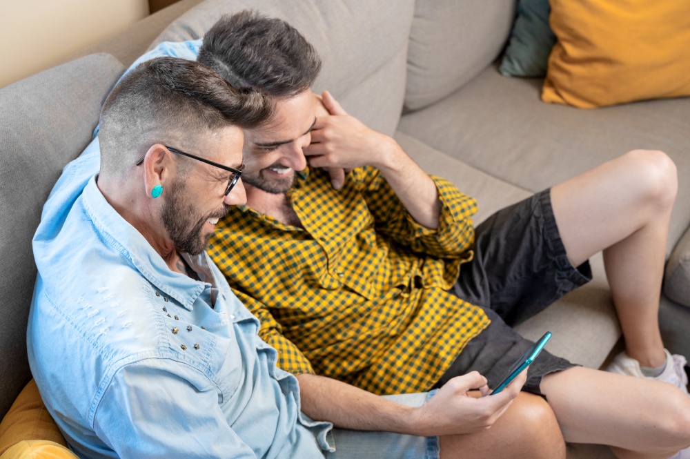Cheerful gay couple sitting on the couch hugging and looking at the mobile phone, watching a movie, scrolling through the social network. High quality photography.. Cheerful gay couple sitting on the couch hugging and looking at the mobile phone, watching a movie, scrolling through the social network