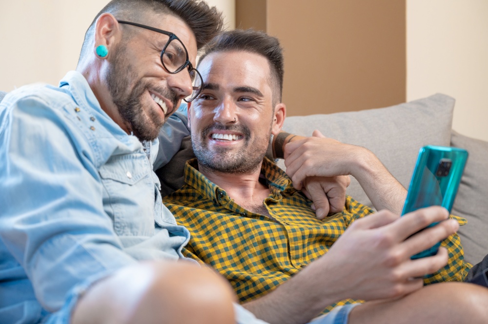 Cheerful gay couple sitting on the couch hugging and looking at the mobile phone, watching a movie, scrolling through the social network. High quality photography.. Cheerful gay couple sitting on the couch hugging and looking at the mobile phone, watching a movie, scrolling through the social network