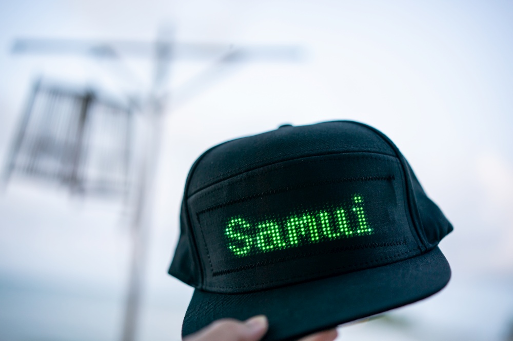 Hand holding black baseball cap with LED letter "Samui", summer vacation.