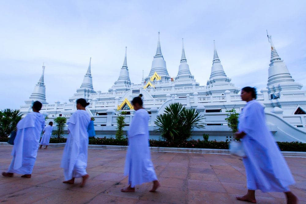 A group of Buddhist women in white traditional clothing are practices Dharma in front of the white pagodas in Uposatha Day. Wat Asokaram, Thailand. Motion blur. Focus on pagodas.