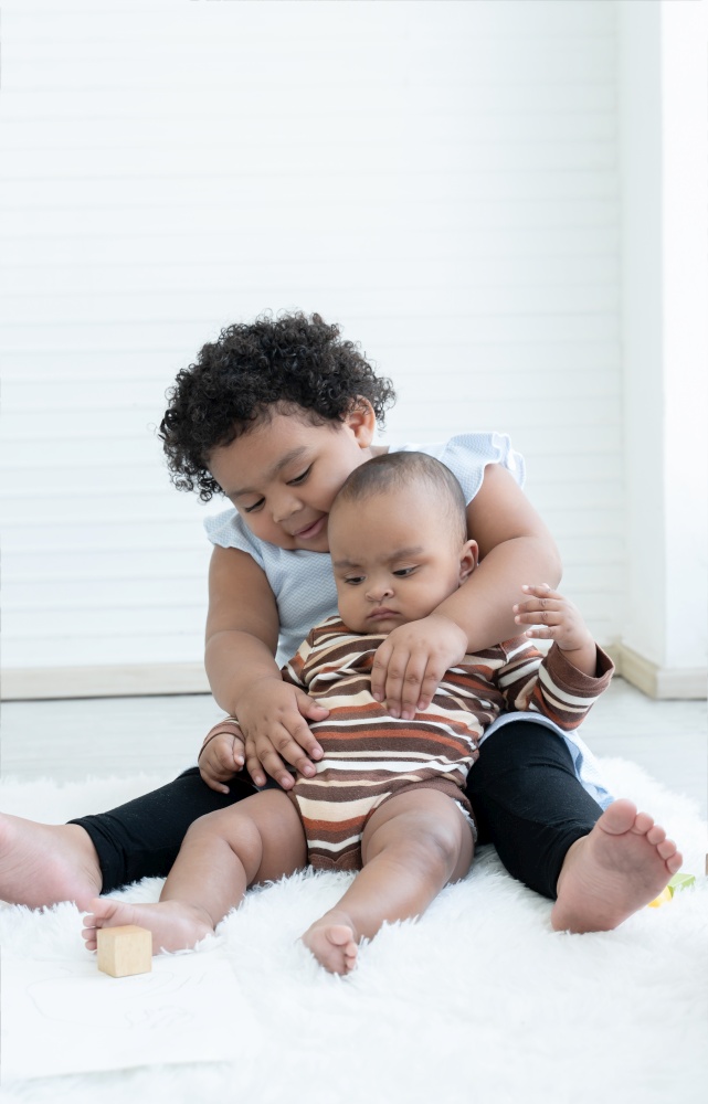 Happy Little cute African chubby kid girl hugging her newborn baby sister in arms while sitting playing together on floor at home. Siblings bond relationship concept. White background