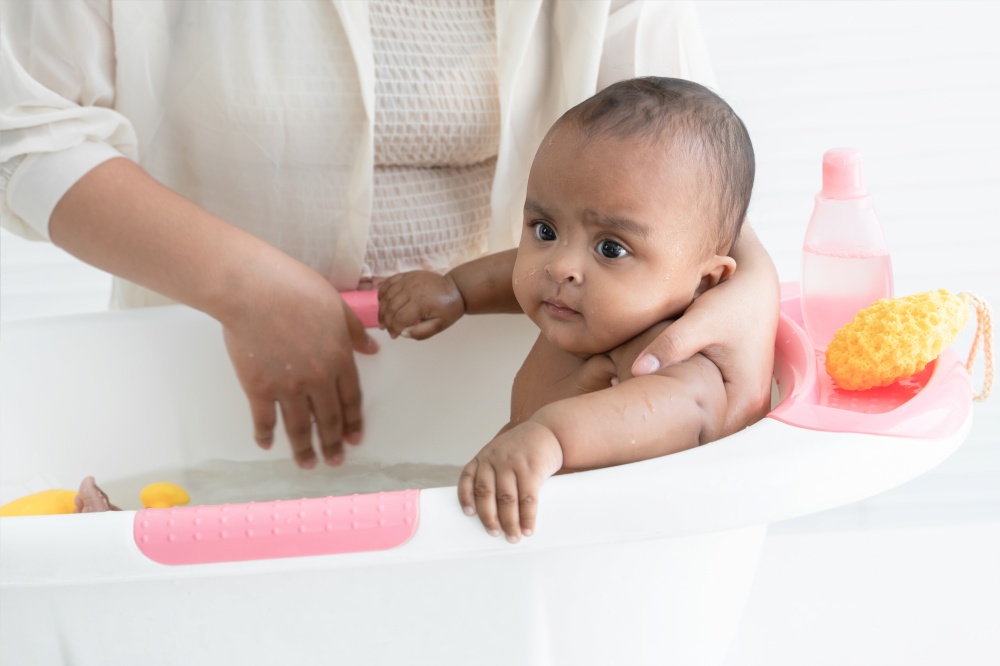 Happy adorable African newborn baby bathing in bathtub. Mother washing her little daughter in warm water with yellow sponge and shower gel. Newborn baby cleanliness care concept