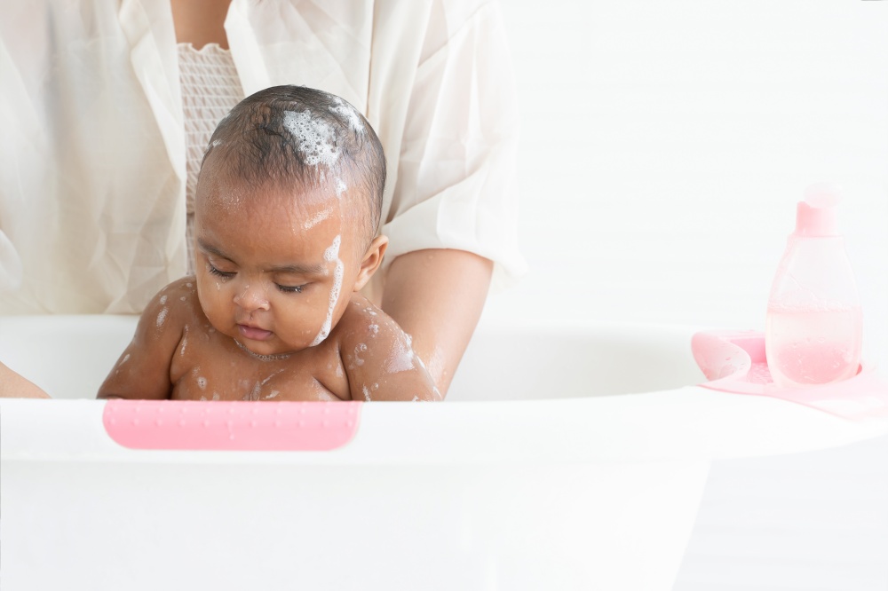 Cute African newborn baby bathing in bathtub with soap bubbles on head and body. Mother washing her little daughter in warm water. Newborn baby cleanliness care concept