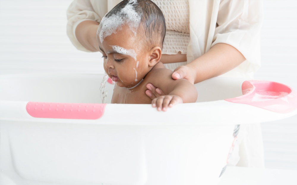 Cute African newborn baby bathing in bathtub with soap bubbles on head and body. Asian young mother washing her little daughter in warm water. Newborn baby cleanliness care concept