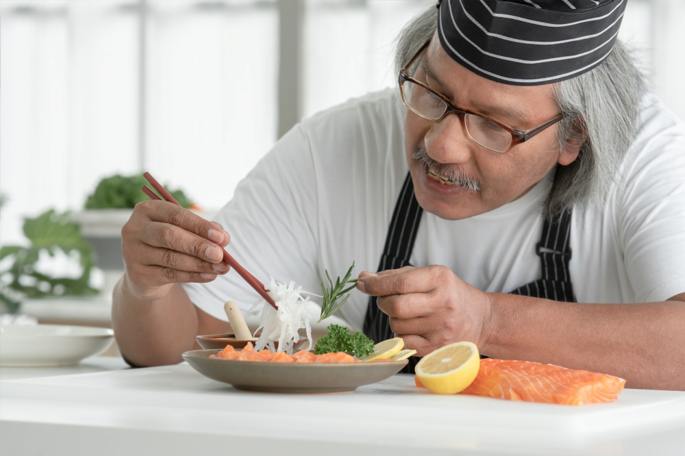 Happy Asian senior chef man holding chopsticks to meticulous arrange radish, thyme, parsley and lemon on sliced fresh salmon sashimi in plate ready to serve. Japanese food home cooked concept