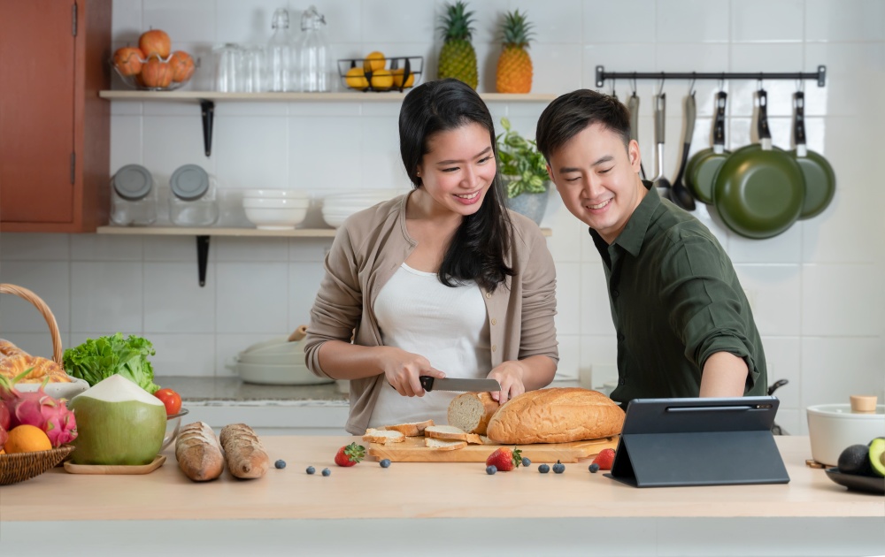 Young Asian romantic couple is cooking in the kitchen. A beautiful woman and handsome man making healthy food while looking recipe and cooking steps on tablet together