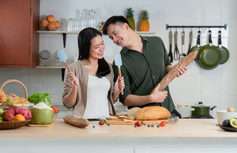Young Asian couple having fun in kitchen, singing and dancing while cooking at home. Playful lover using spatulas as microphones and loaf stick of bread as guitar doing band together