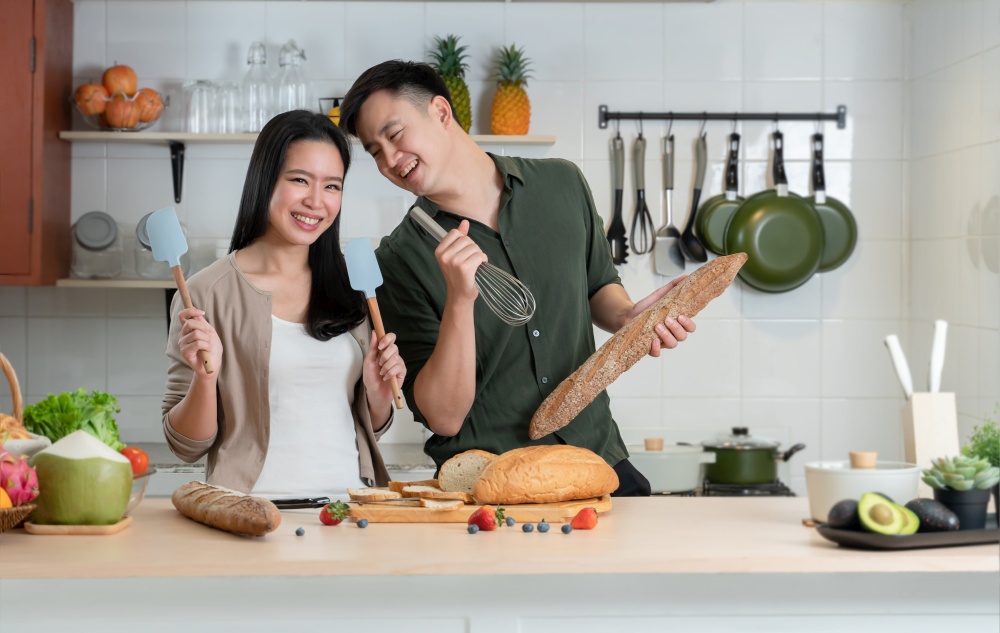 Young Asian couple having fun in kitchen, singing and dancing while cooking at home. Playful lover using spatulas and egg whisk as microphones and loaf stick of bread as guitar doing band together
