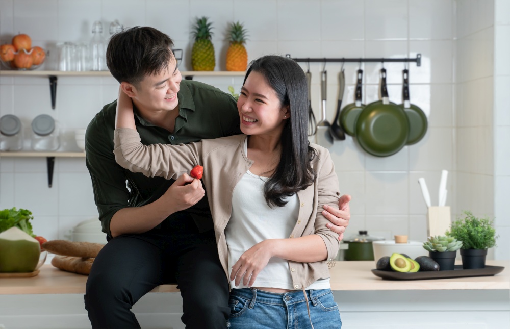 Portrait of young Asian romantic couple standing in the kitchen, happy lover hugging, smiling and looking at each other, handsome man feeding strawberry to beautiful woman while cooking together