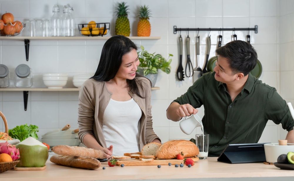 Young Asian romantic couple is cooking in the kitchen. A beautiful woman and handsome man making healthy breakfast food while boyfriend pouring milk into glass and smile at his girlfriend