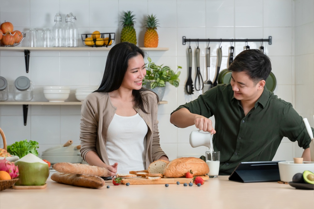 Young Asian romantic couple is cooking in the kitchen. A beautiful woman and handsome man making healthy breakfast food while boyfriend pouring milk into glass for his girlfriend