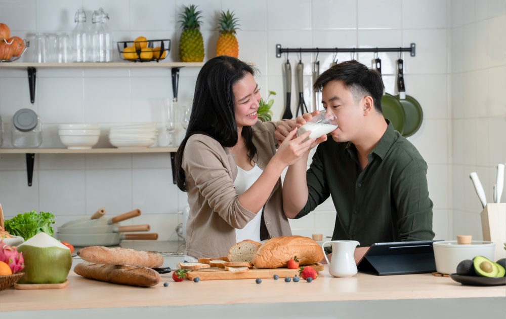 Young Asian romantic couple is cooking in the kitchen. Happy beautiful woman holding milk of glass and feeding milk to her handsome man while making healthy breakfast food at home together