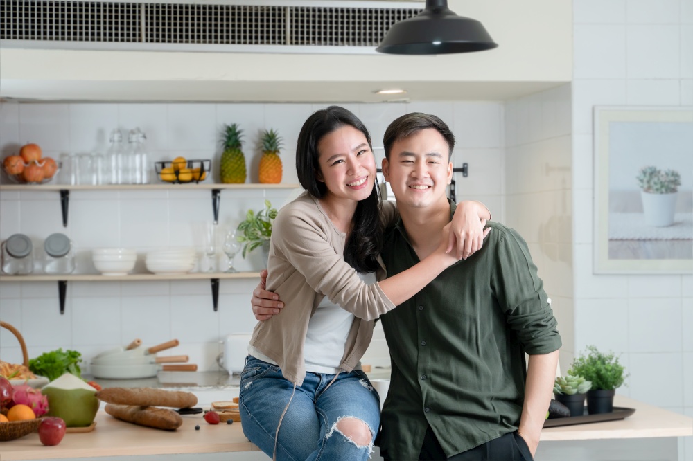 Portrait of young Asian couple standing in the kitchen, happy romantic lover smiling and looking at each other, beautiful woman and handsome man hugging while cooking together at home