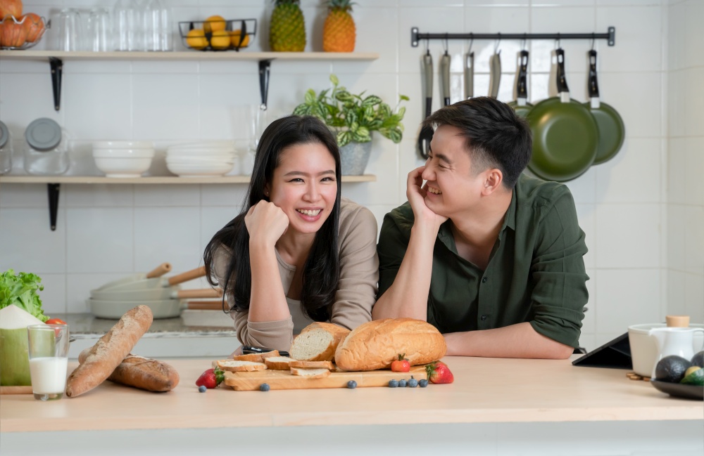 Portrait of young Asian romantic couple smiling, placing hands on chin in the kitchen, happy beautiful wife and handsome husband cooking healthy breakfast food together at home