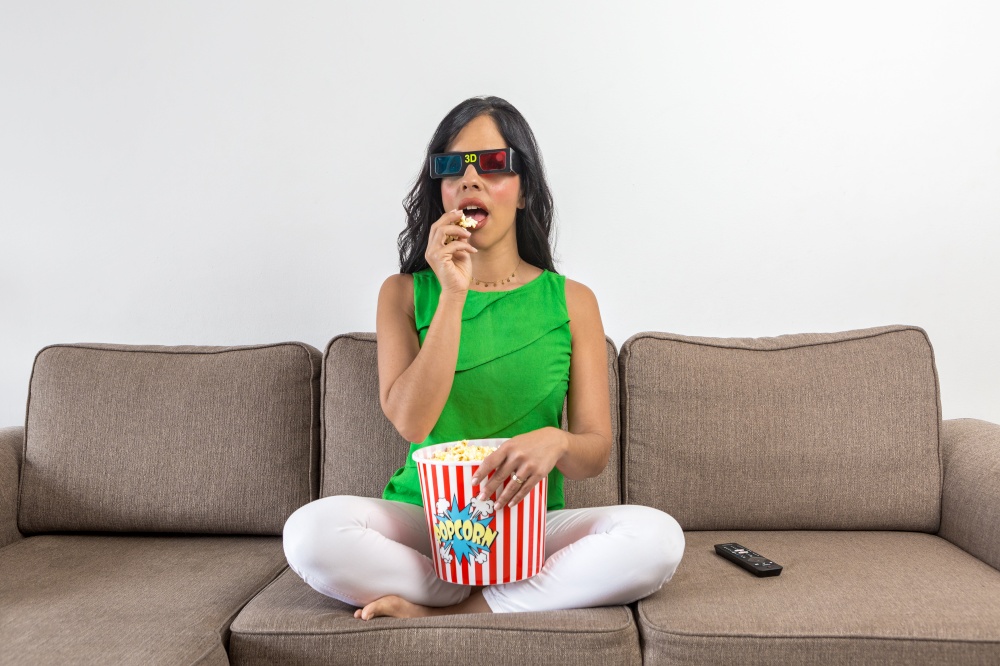 Hispanic female in casual clothes and 3D glasses eating fresh popcorn and watching film on TV while sitting cross legged on sofa in daytime at home. Woman eating popcorn during showtime