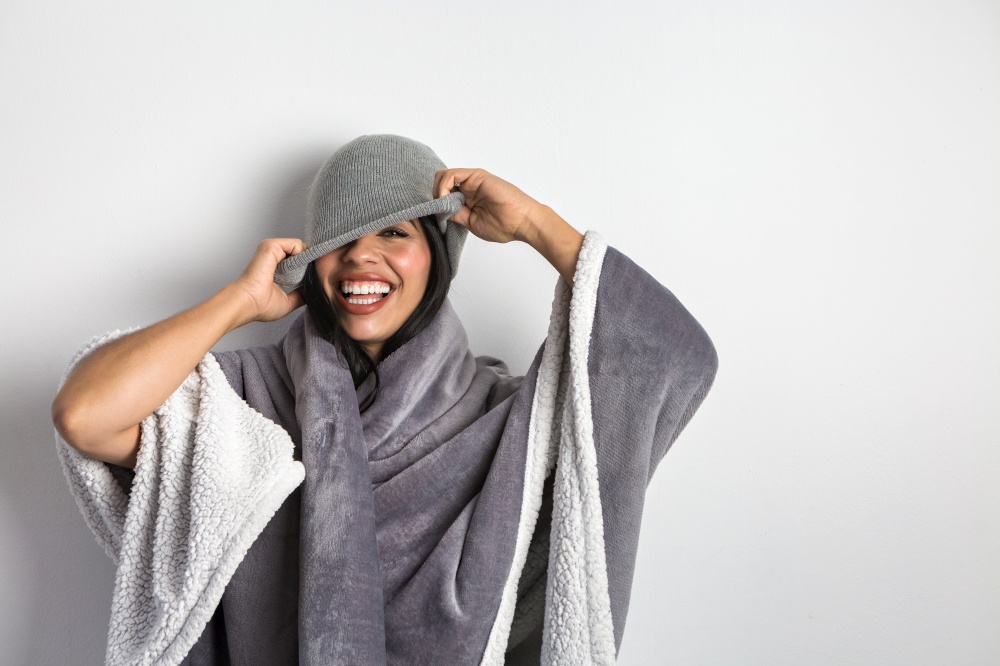 Content Hispanic female model wrapped in soft blanket laughing happily while covering eye with beanie against white background. Cheerful ethnic woman in blanket taking on gray hat