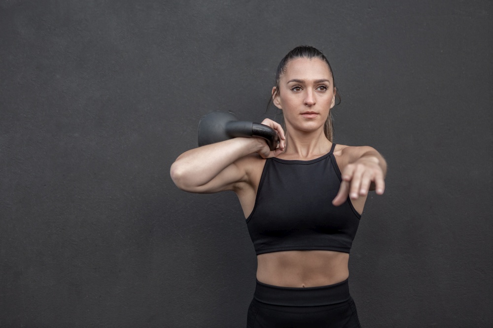 Fit female athlete in sportswear stretching out arm and holding heavy kettlebell on shoulder during functional training against black wall in gym. Determined sportswoman exercising with kettlebell