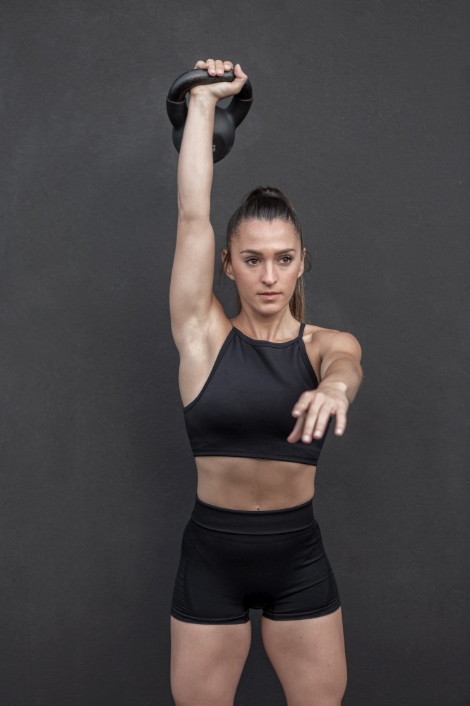 Muscular sportswoman in activewear raising arm with heavy kettlebell over head and looking away against black background during intense workout in gym. Strong female athlete lifting kettlebell
