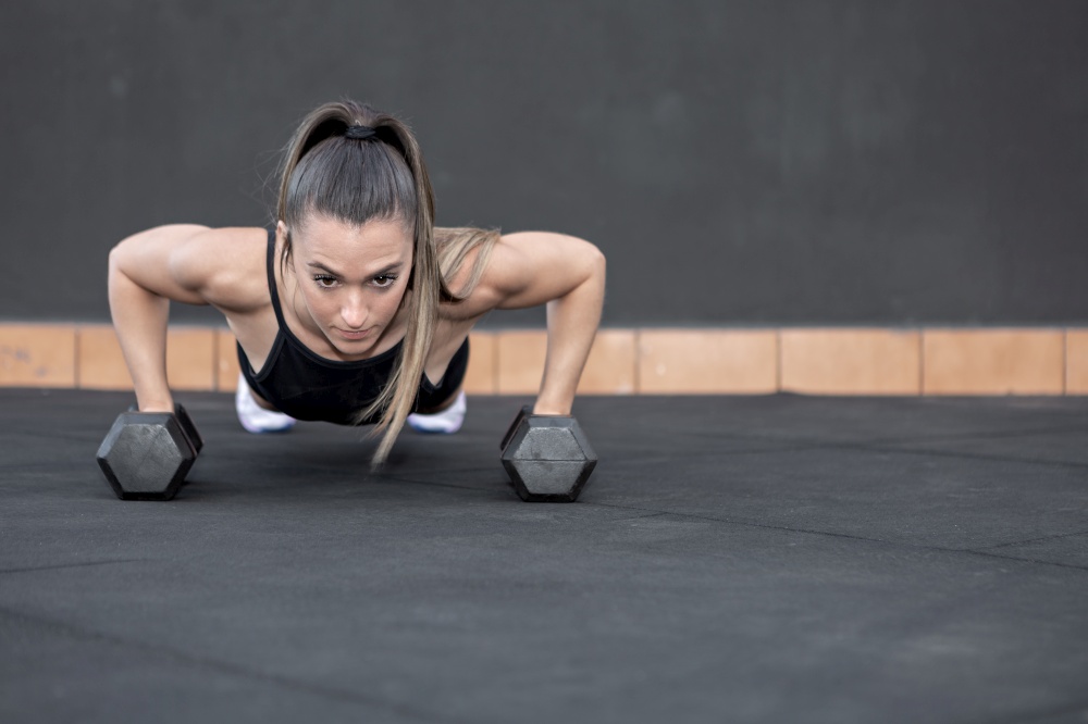 Ground level of determined female athlete with ponytail doing push up exercise on dumbbells during functional workout in gym. Sportswoman doing push ups on dumbbells
