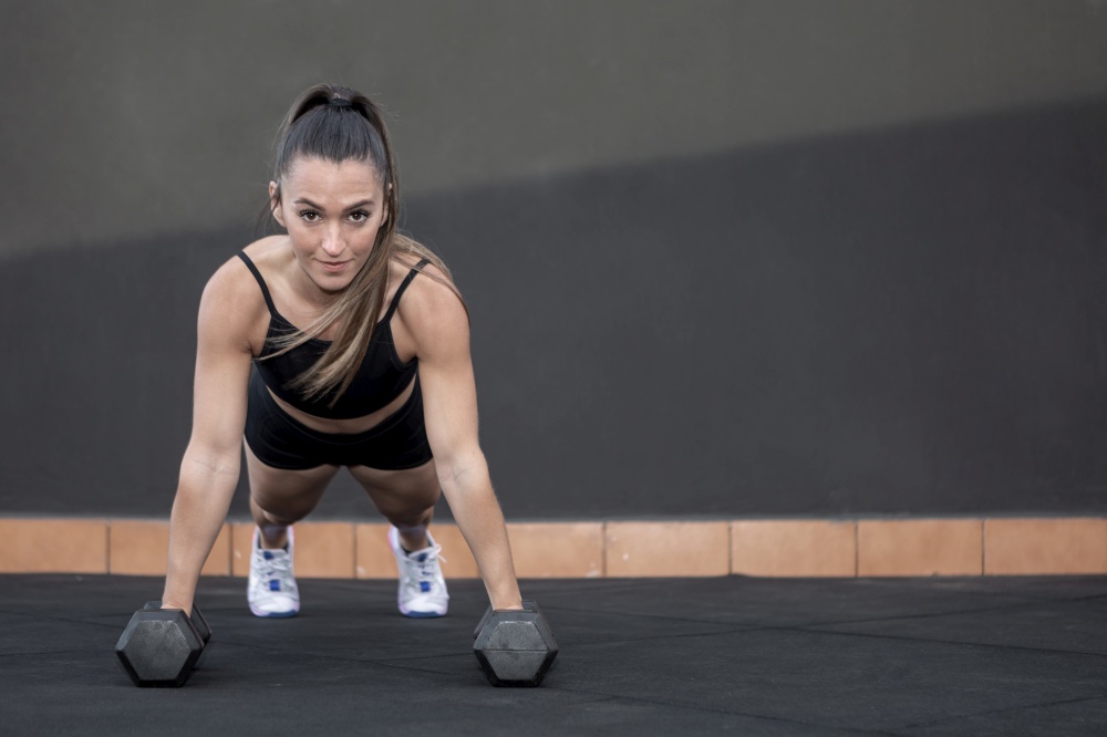 Fit female athlete in sportswear with ponytail doing plank exercise on dumbbells and looking at camera against black wall during fitness workout in gym. Sportswoman doing plank on dumbbells