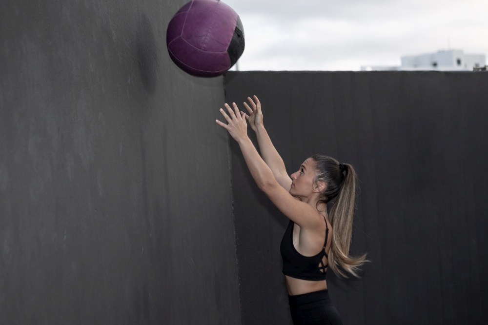 Side view of female athlete with ponytail throwing heavy ball at black wall during intense fitness workout on sports ground. Sportswoman tossing medicine ball at wall