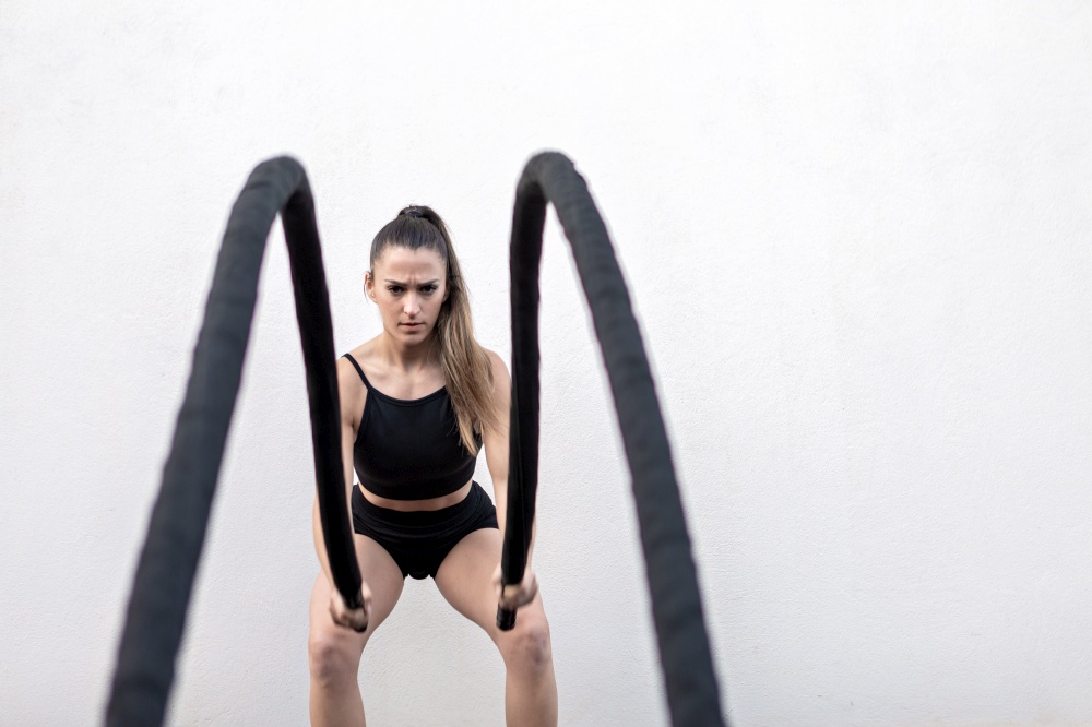 Determined female athlete in black sportswear with ponytail frowning and doing exercise with battle ropes against white wall during intense workout in gym. Sportswoman exercising with battle ropes