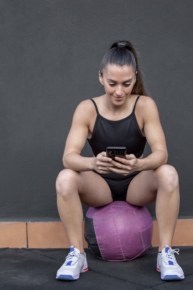 Positive female athlete in sportswear with ponytail sitting on ball against black wall and browsing cellphone during break in fitness workout in gym. Sportswoman using smartphone during break