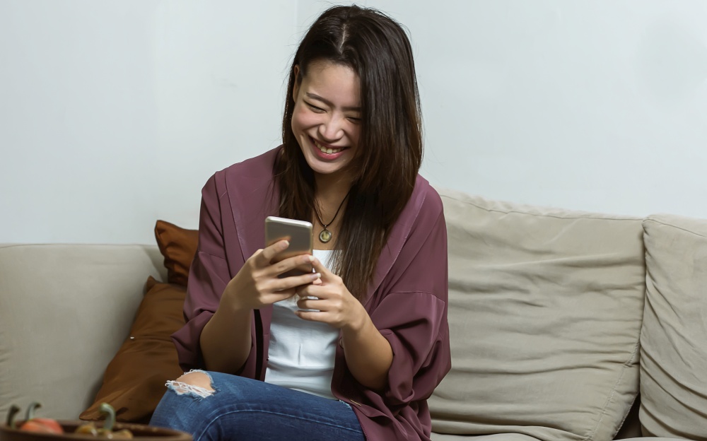 Asian young beautiful woman smiling and chatting by using mobile phone  while relaxing in living room at home. Lifestyle, Technology and New Normal Concept.