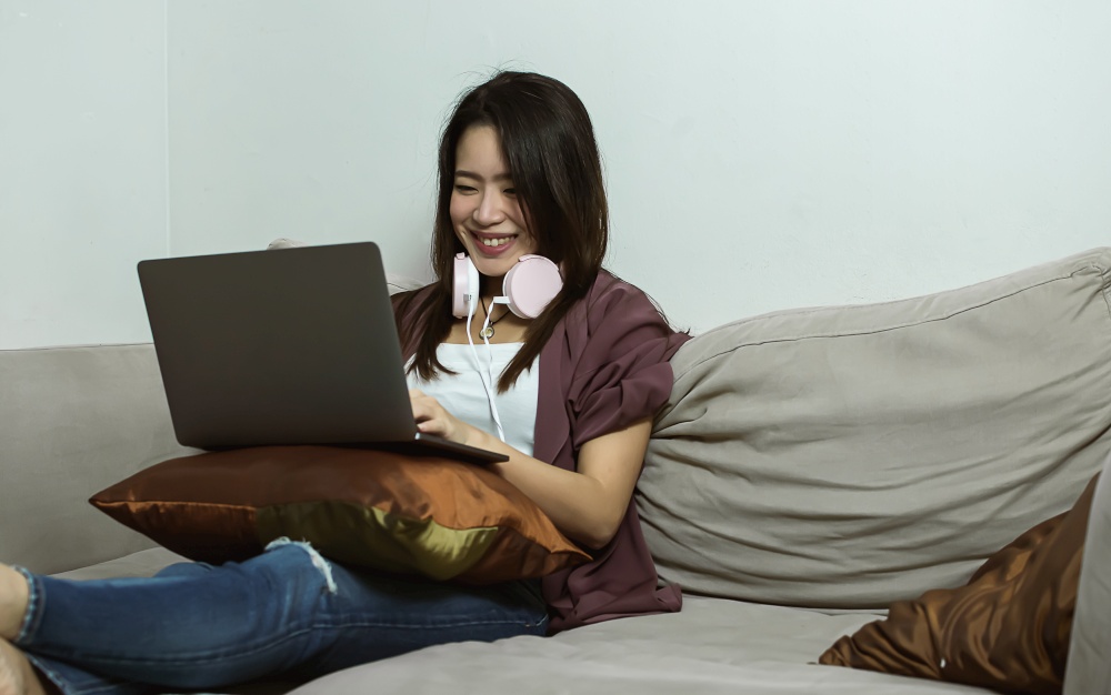 Asian young beautiful woman using laptop and headphone while relaxing in living room at home. Lifestyle, Technology and New Normal Concept.