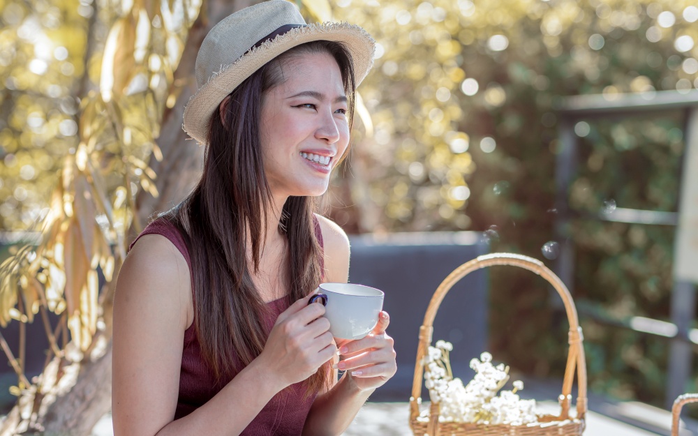 Asian beautiful woman wearing hat, smiling and drinking tea in the garden. Lifestyle, Summer and Travel Concept.