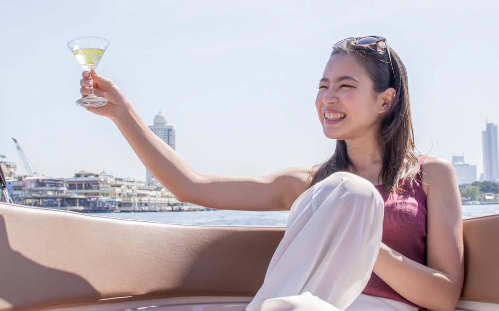 Asian beautiful woman drinking white wine and making toast while traveling and sitting on boat with background of beautiful building and landscape. Summer Travel and Lifestyle Concept.