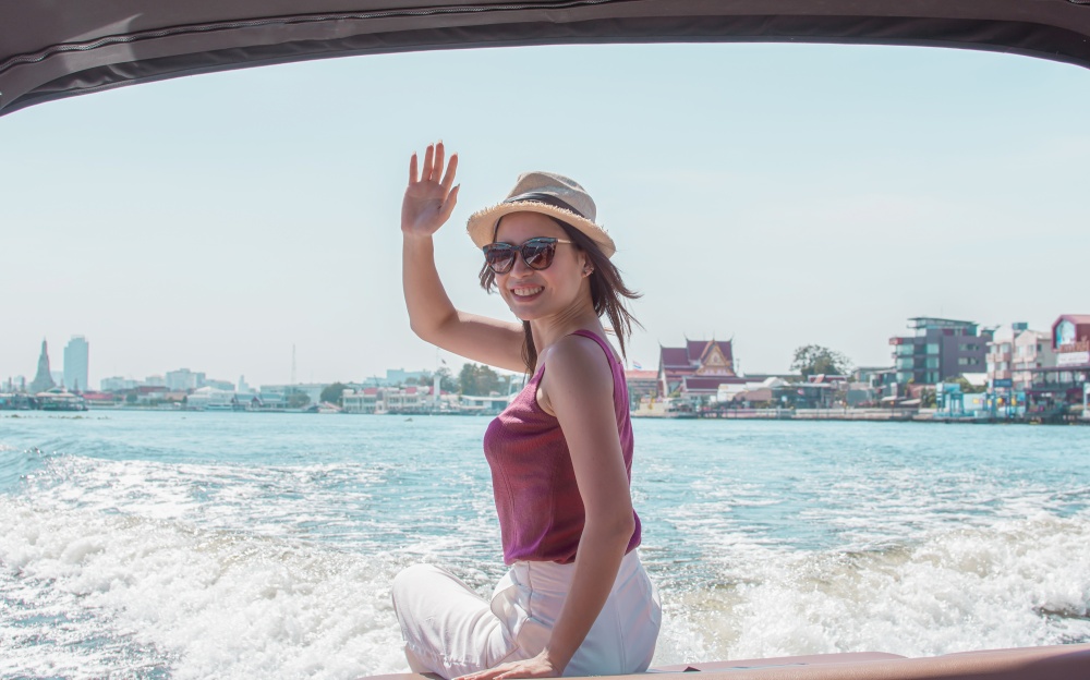 Asian beautiful woman wearing hat and sunglasses, waving her hand while sitting on boat with background of beautiful landscape. Lifestyle and Travel Concept.