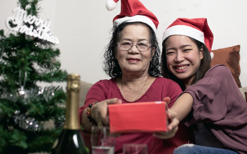 Two 60s old mother and daughter wearing santa hat, hug and holding gift at home with blur foreground of tree and bottle. Christmas or New Year Celebration Concept.